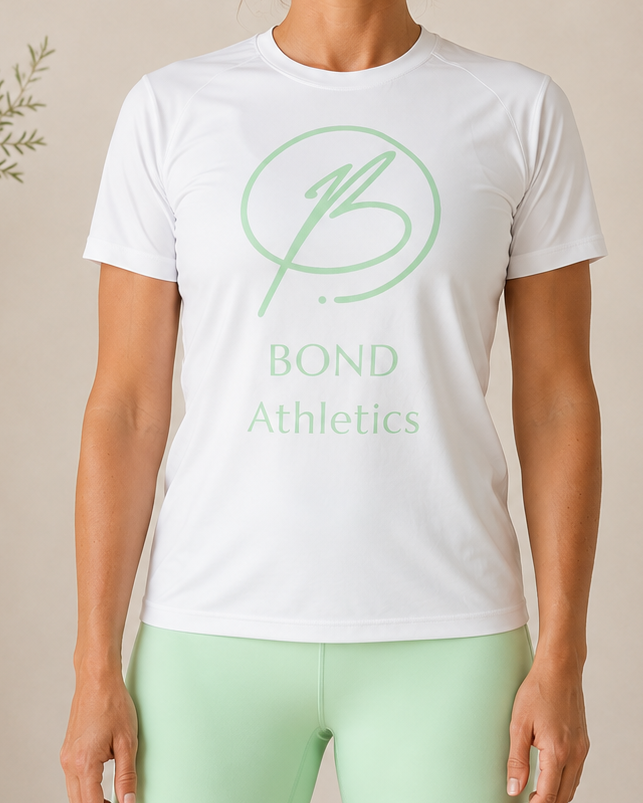 A woman stands indoors wearing a white short-sleeve athletic T-shirt with a light green circular logo and the words 'BOND Athletics' on the front, paired with mint green leggings. The background is a warm beige wall with soft decorative plants and furniture partially visible.