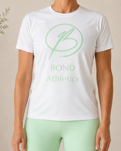 A woman stands indoors wearing a white short-sleeve athletic T-shirt with a light green circular logo and the words 'BOND Athletics' on the front, paired with mint green leggings. The background is a warm beige wall with soft decorative plants and furniture partially visible.