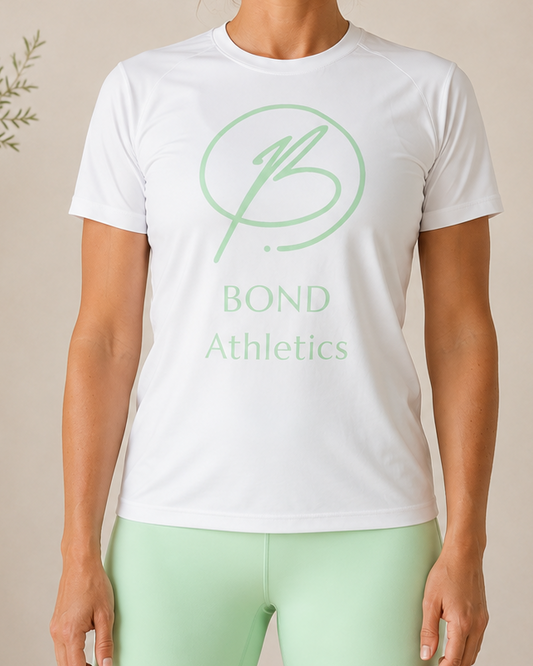 A woman stands indoors wearing a white short-sleeve athletic T-shirt with a light green circular logo and the words 'BOND Athletics' on the front, paired with mint green leggings. The background is a warm beige wall with soft decorative plants and furniture partially visible.