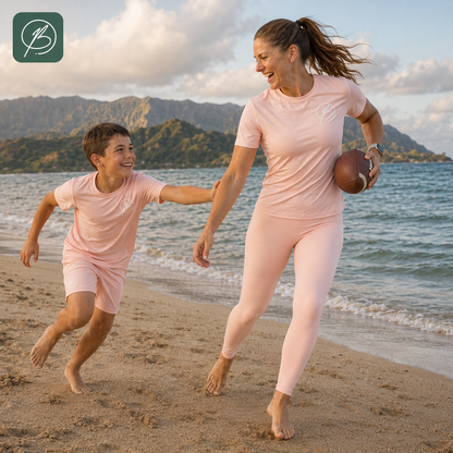A woman and a boy running hand in hand on a sandy beach, wearing matching light pink athletic sets. The woman holds a football, with ocean water and mountains in the background.