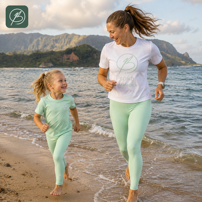 A woman and a young girl running on a sandy beach at the water's edge, wearing matching mint green athletic sets. The woman wears a white t-shirt with a green 'Bond Athletics' logo and mint green leggings, while the girl wears a matching mint green t-shirt and leggings.