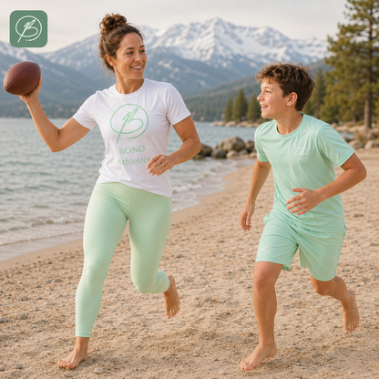 A woman and a boy running on a sandy beach with water and mountains in the background. The woman wears a white Bond Athletic t-shirt and mint green leggings, holding a football, while the boy wears a matching mint green t-shirt and shorts.
