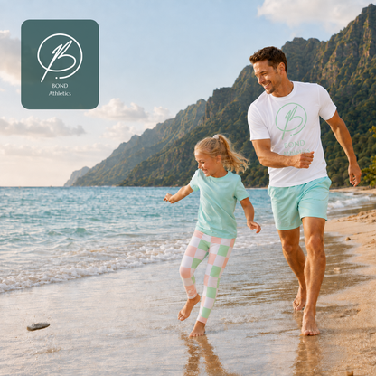 A man and a young girl running on a sandy beach with ocean waves and green mountains in the background. The man wears a white athletic T-shirt and mint green shorts, and the girl wears a mint green T-shirt and pastel pink and mint checkered leggings.