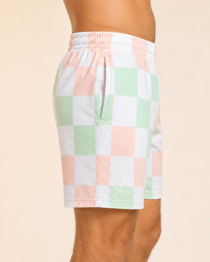 A side profile view of a person wearing checkered shorts with a pastel checkerboard pattern of pink, mint green, and white, set against a solid light beige background.