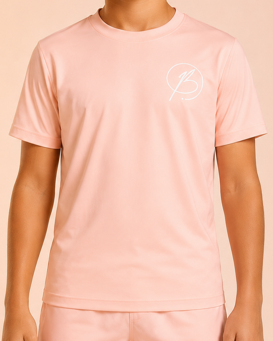 A child wearing a light pink short-sleeve t-shirt and matching shorts set with a small white circular logo on the chest and left thigh, set against a solid peach-pink background.