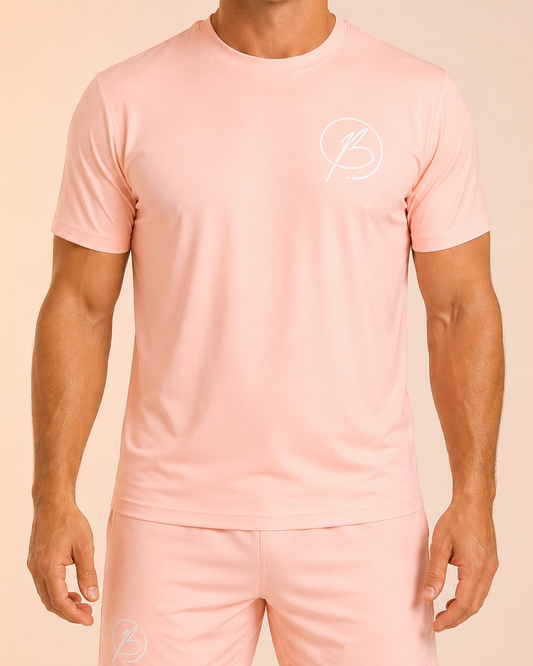A man wearing a light pink athletic t-shirt and matching shorts with a small white circular logo on the chest and left thigh, set against a solid pale peach background.