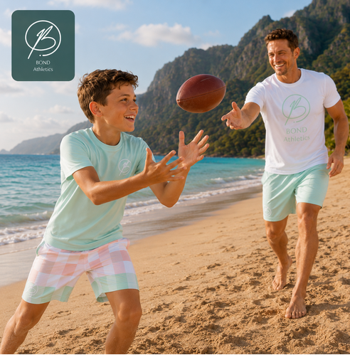 A father and son playing with a football on a sunny beach, both wearing matching light mint green t-shirts and shorts from Bond Athletics. A dark green logo badge for Bond Athletics is in the top-left corner.