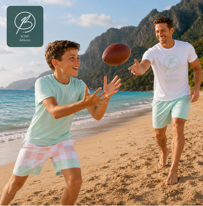 A father and son playing with a football on a sunny beach, both wearing matching light mint green t-shirts and shorts from Bond Athletics. A dark green logo badge for Bond Athletics is in the top-left corner.