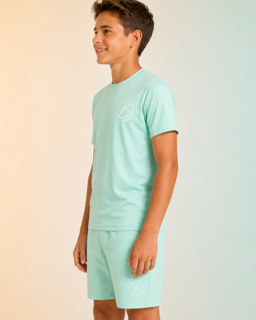 A young boy wearing a mint green short-sleeve t-shirt and matching shorts with a small white circular logo on the chest and thigh, standing against a split beige and light blue background.