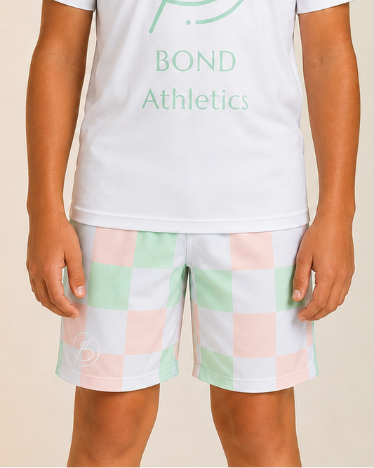 A child model wearing a white short-sleeve t-shirt with a mint green 'Bond Athletics' logo and checkered athletic shorts in pastel pink, mint green, and white, set against a soft beige-to-green gradient background.