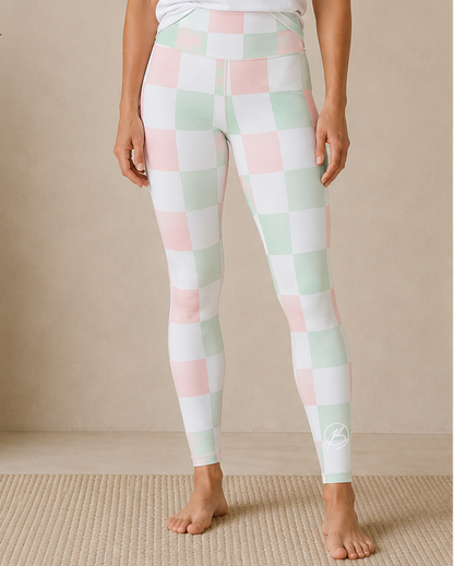 A woman stands barefoot indoors wearing a white short-sleeve athletic T-shirt with a mint green 'BOND Athletics' logo and pastel checkered leggings in white, pale pink, and mint green.