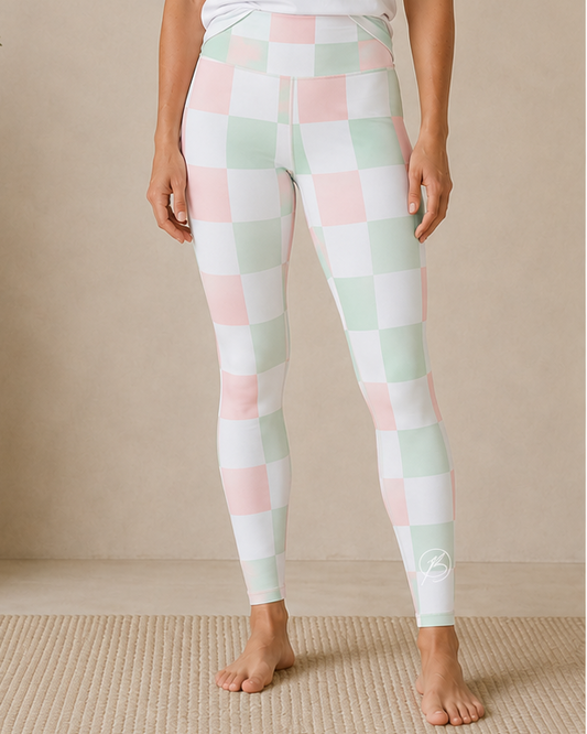 A woman stands barefoot indoors wearing a white short-sleeve athletic T-shirt with a mint green 'BOND Athletics' logo and pastel checkered leggings in white, pale pink, and mint green.