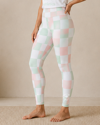 A woman stands barefoot indoors wearing white athletic leggings with a pastel pink, mint green, and white checkerboard pattern, paired with a white fitted T-shirt. The room has a beige wall, a large vase with dried branches, and a small wooden bench with a folded towel.