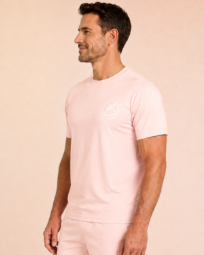 A man wearing a light pink short-sleeve t-shirt and matching shorts with a small white circular logo on the chest and thigh, standing against a solid pale peach background.