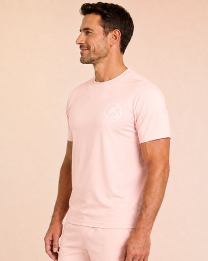 A man wearing a light pink short-sleeve t-shirt and matching shorts with a small white circular logo on the chest and thigh, standing against a solid pale peach background.