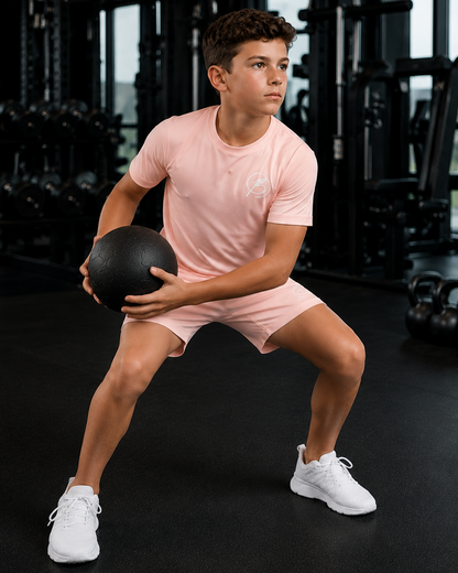A young man in a pink athletic t-shirt and matching shorts holds a black medicine ball in a gym setting.