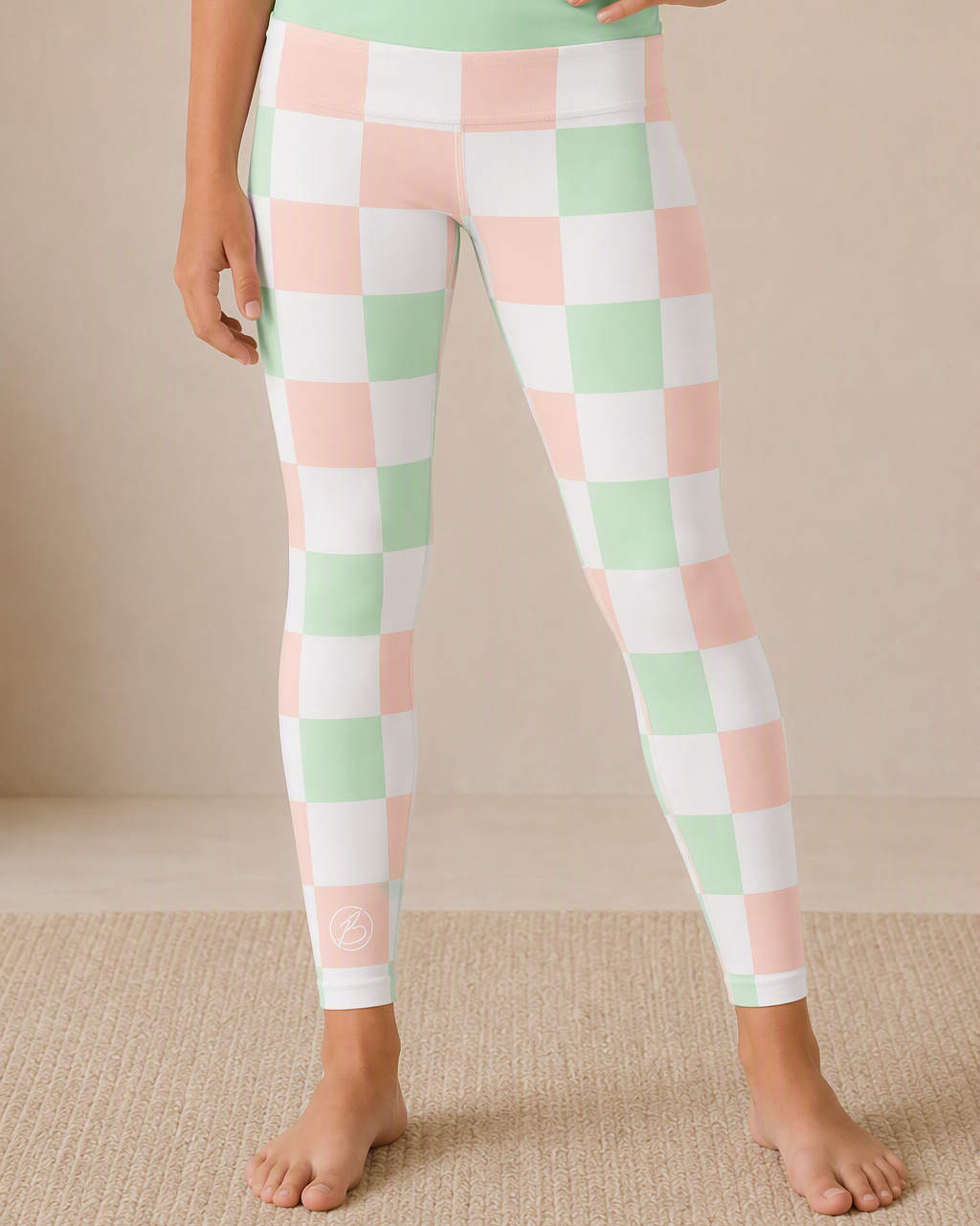 A person wearing checkered leggings in pastel pink, mint green, and white, standing barefoot on a beige textured rug against a neutral beige wall.