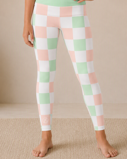 A person wearing checkered leggings in pastel pink, mint green, and white, standing barefoot on a beige textured rug against a neutral beige wall.