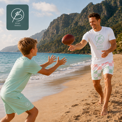 A man and a boy play catch with a football on a sandy beach with ocean and mountains in the background. Both are wearing matching light mint green Bond Athletics t-shirts and shorts sets.