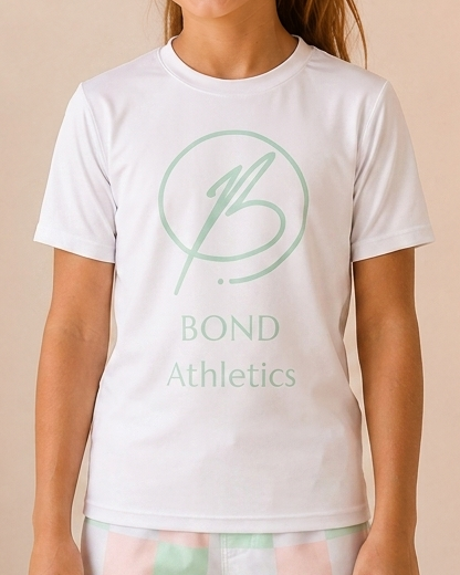 A young girl stands in a studio setting wearing a white t-shirt with a mint green 'Bond Athletics' logo and pastel checkered leggings. The background features a warm beige wall, a large textured vase with dried branches, and a wooden stool with a folded blanket.