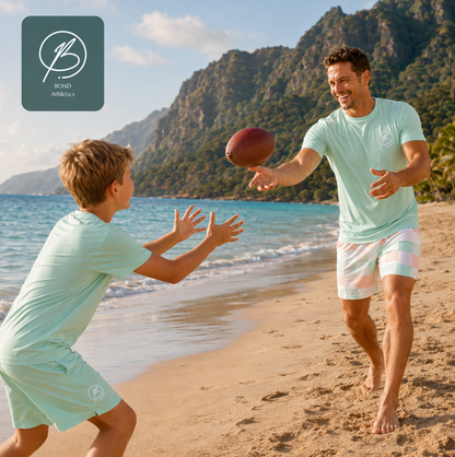A father and son playing with a football on a sandy beach, both wearing matching mint green athletic t-shirts and shorts. The father is wearing the brand's shorts, and a logo overlay for 'BOND Athletics' is visible in the top left corner.