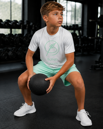 A young boy in a gym squatting while holding a black medicine ball, wearing a white t-shirt with a green circular logo that reads 'BOND MAGIC' and mint green athletic shorts.