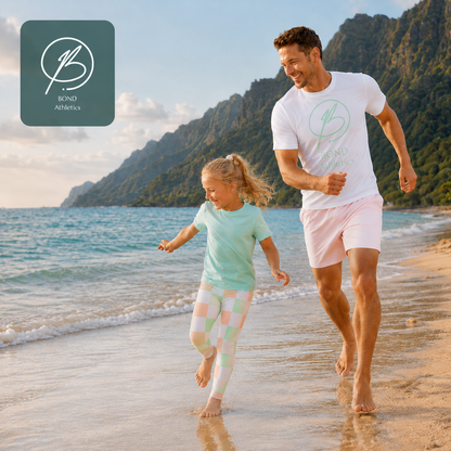 A man and a young girl run happily along a sandy beach with ocean waves and green mountains in the background. Both are wearing matching white Bond Athletics t-shirts with a light green logo and Bond Kids leggings.