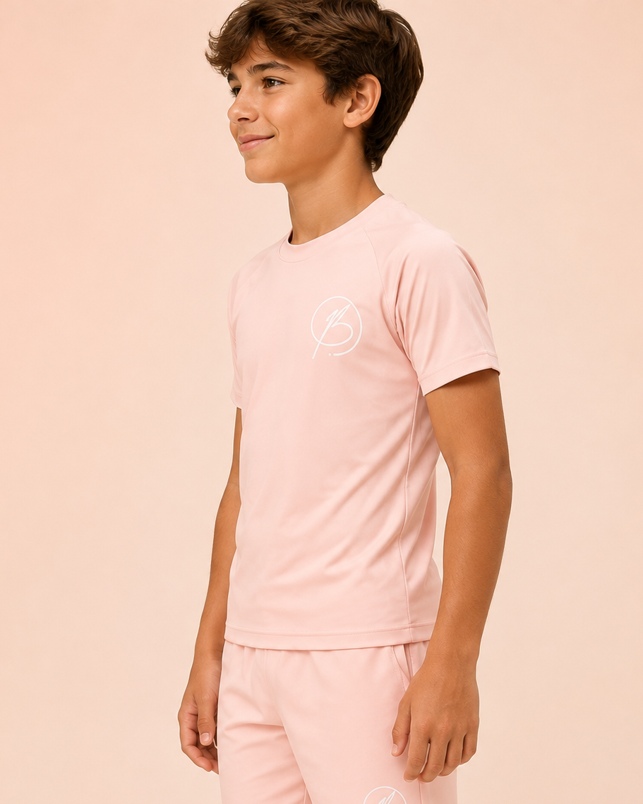 A young boy modeling a light pink short-sleeve t-shirt and matching shorts set, featuring a small white circular logo on the chest and thigh, set against a solid pale peach background.